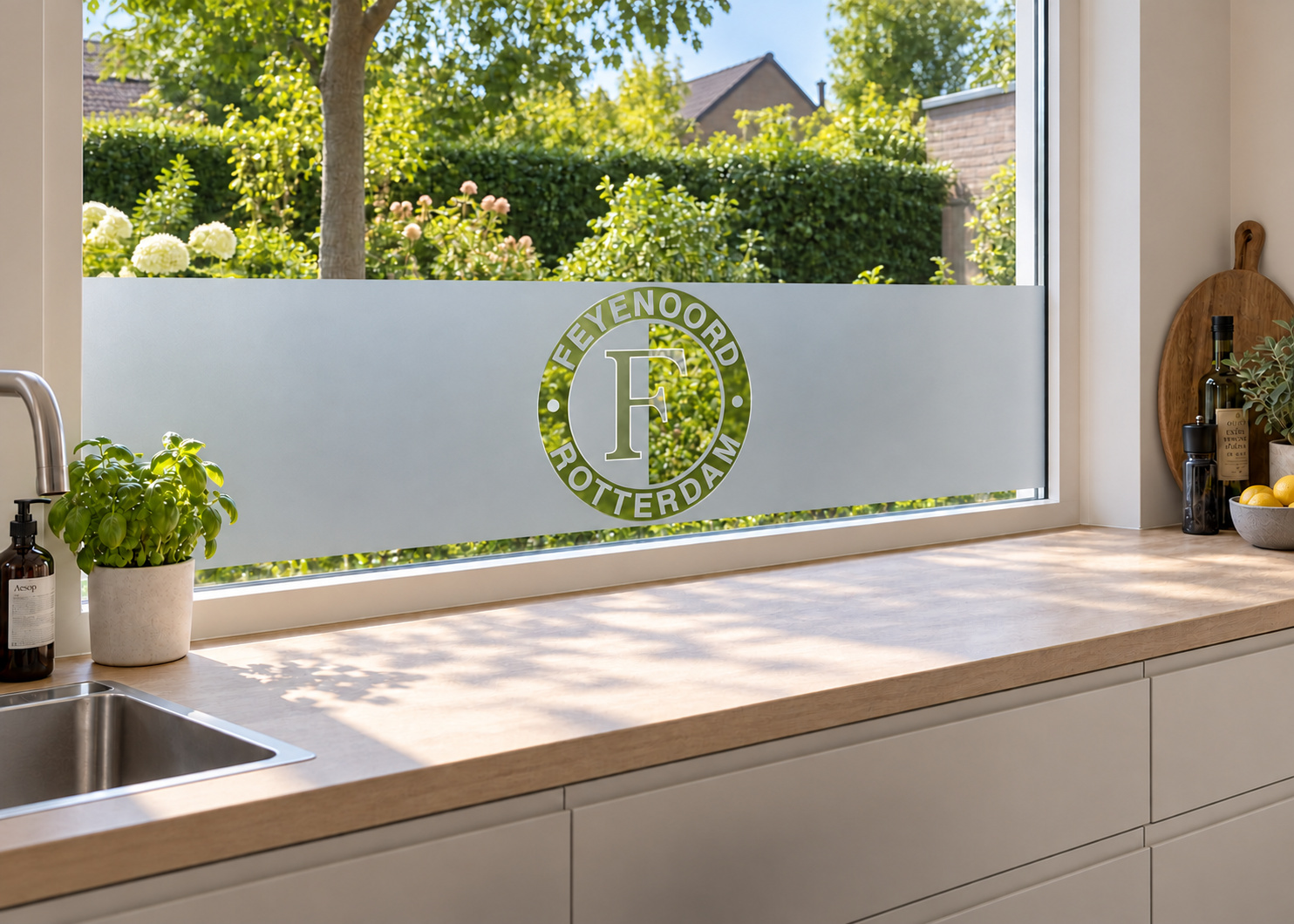 Raamfolie Feyenoord logo (RFH039) als horizontale raamfolie boven het aanrecht in een keukenraam, met matte folie en doorkijk naar de tuin.